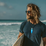 Surfbrett-Anhänger aus Maserholz und blau-weißem Epoxidharz, hochglanzpoliert, ca. 6,5 × 2 cm. An schwarzer Lederkette mit Edelstahl-Karabiner und 5-cm-Verlängerung; Edelstahl-Öse. Im Tageslicht lagunenblau, unter Schwarzlicht fluoreszierend – ideal als Party-/Festival-Schmuck. Handgefertigtes Unikat, Lieferung in edler Schmuckbox.