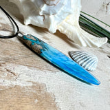 Surfbrett-Anhänger aus Maserholz und blau-weißem Epoxidharz, hochglanzpoliert, ca. 6,5 × 2 cm. An schwarzer Lederkette mit Edelstahl-Karabiner und 5-cm-Verlängerung; Edelstahl-Öse. Im Tageslicht lagunenblau, unter Schwarzlicht fluoreszierend – ideal als Party-/Festival-Schmuck. Handgefertigtes Unikat, Lieferung in edler Schmuckbox.