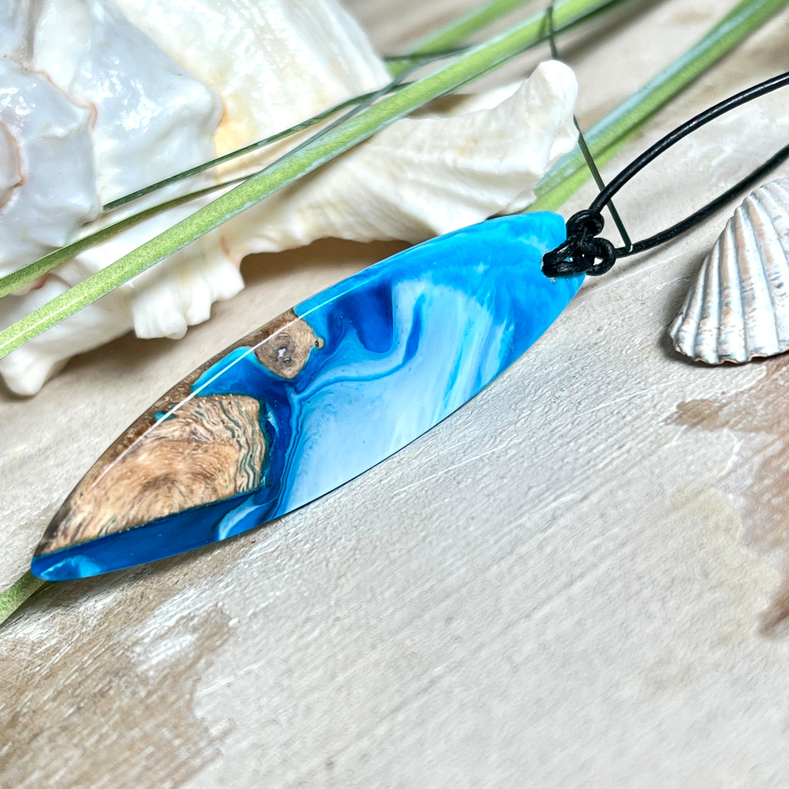 Handgefertigter Surfbrett-Anhänger „Ocean Glide“ aus Maserholz und Epoxidharz in verschiedenen Blau-, und Weißtönen. Der ca. 6,5 × 2 cm große Anhänger wird an einem verstellbaren Lederband (26–46 cm) mit Holzperlen geliefert. Ein maritimes Unikat mit natürlicher Holzmaserung, das die Schönheit des Meeres einfängt.
