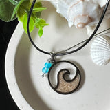 Wave Spirit – runder Edelstahlanhänger mit echter Sandfüllung in Harz eingebettet, inspiriert von einer stilisierten Welle. Verziert mit zwei blauen Achatperlen. Der Anhänger (ca. 2,5 cm Durchmesser) hängt an einer 45 cm langen Lederkette mit Edelstahlverschluss und 5 cm Verlängerungskette. Maritimer Look trifft moderne Eleganz – ein handgefertigtes Schmuckstück für Meerliebhaber