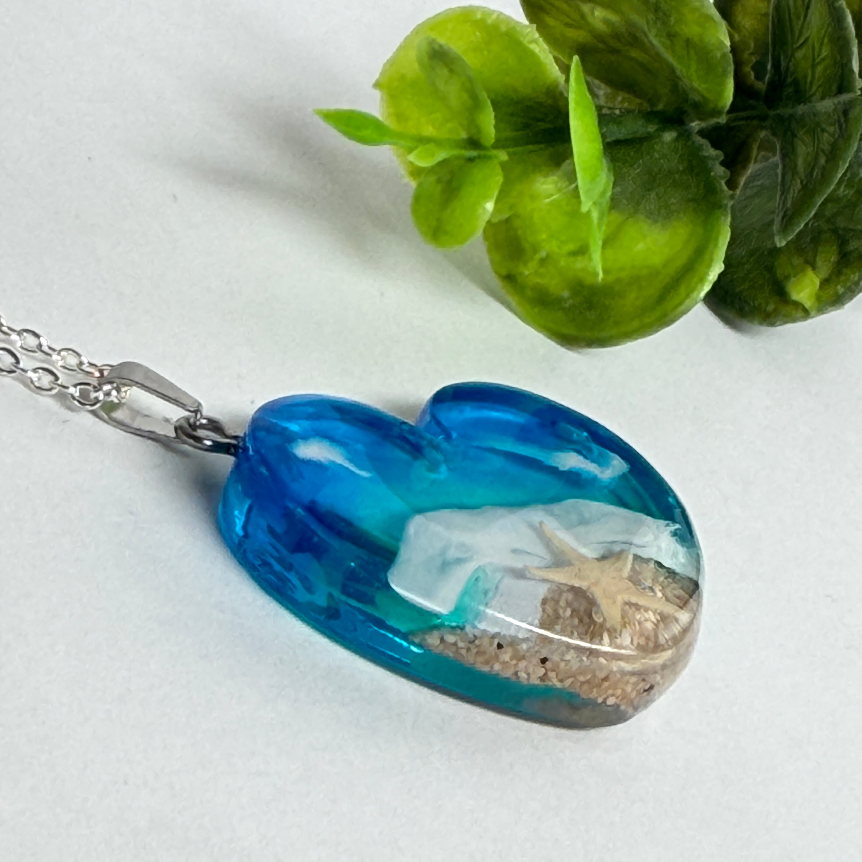 Ein herzförmiger Anhänger mit einer ganzen Ozeanwelt darin: Welle, Sand und Seestern in leuchtendem Blau und Türkis – handgegossen, einzigartig, unvergesslich. Mit versilberter Kette (46 cm) und Edelstahl-Karabiner.