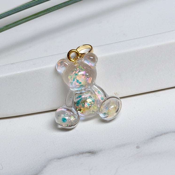 Kleiner Bärchen-Anhänger aus transparentem Harz mit irisierenden Flakes in Regenbogen-Schimmer, goldfarbene Anhängerschlaufe aus Edelstahl. Größe ca. 2,2 × 2,1 cm. Handgefertigtes Unikat; Flakes-Anordnung variiert, vereinzelt kleine Luftbläschen möglich.