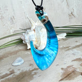 Handgefertigter Haken-Anhänger im Hei-Matau-Stil aus Maserholz und klarem Harz mit Blau-, Türkis- und Weißverlauf, hochglanzpoliert. Oben sichtbares Holz im Harzbereich, getragen an verstellbarem schwarzem Lederband mit Schiebeknoten und Zierperlen. Anhänger ca. 7 × 3 cm, fotografiert mit Muscheln im Hintergrund.