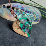 Herzförmiger Anhänger aus Olivenholz mit aquamarinfarbenem Harz und eingelegten Paua-Muschelstücken (Abalone), irisierender Blau-Grün-Schimmer. Der Anhänger hängt an einem braunen, verstellbaren Lederband mit Schiebeknoten; Aufhängung aus Edelstahl. Produktfoto vor maritimer Deko/Abalone-Schale.