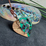 Herzförmiger Anhänger aus Olivenholz mit aquamarinfarbenem Harz und eingelegten Paua-Muschelstücken (Abalone), irisierender Blau-Grün-Schimmer. Der Anhänger hängt an einem braunen, verstellbaren Lederband mit Schiebeknoten; Aufhängung aus Edelstahl. Produktfoto vor maritimer Deko/Abalone-Schale.