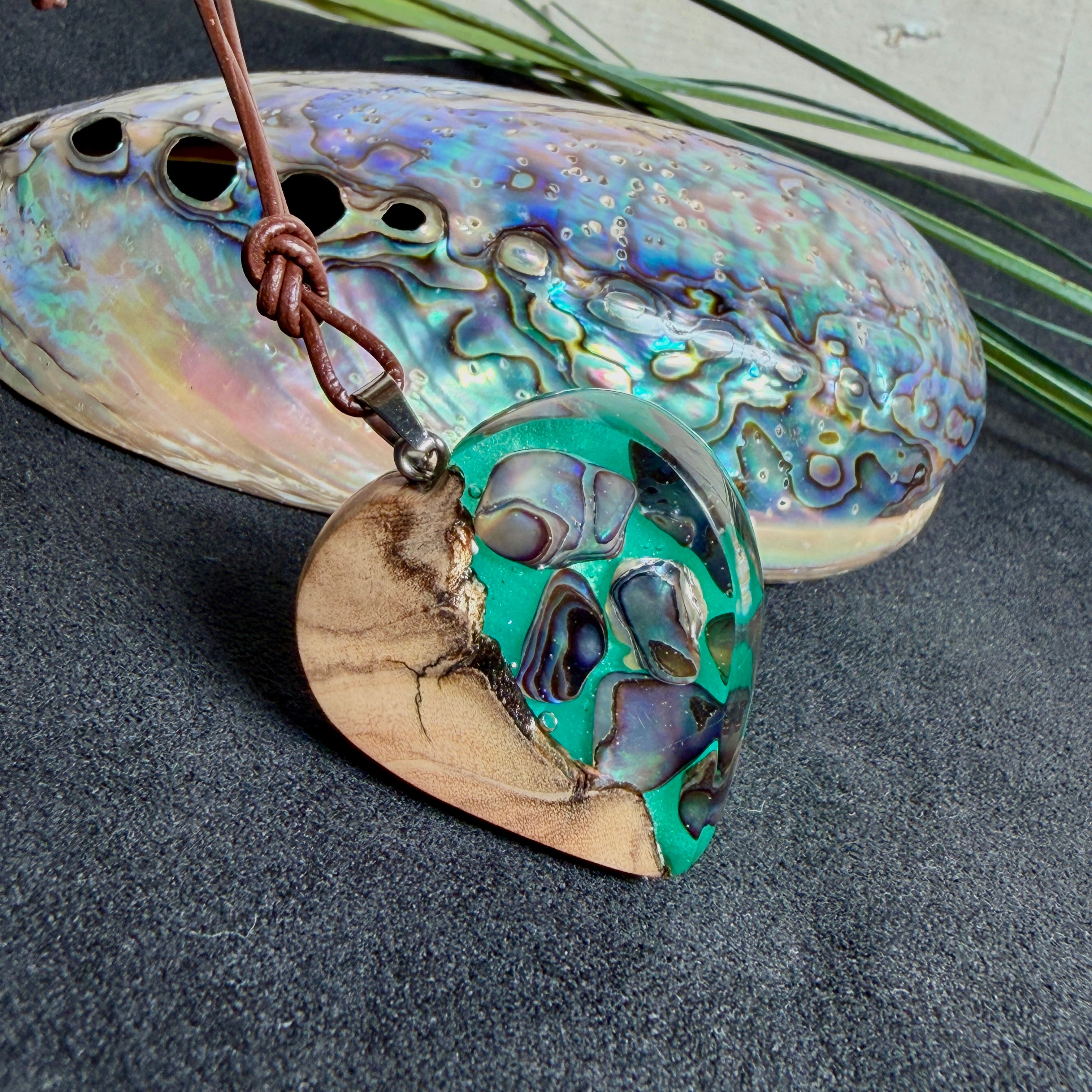 Herzförmiger Anhänger aus Olivenholz mit aquamarinfarbenem Harz und eingelegten Paua-Muschelstücken (Abalone), irisierender Blau-Grün-Schimmer. Der Anhänger hängt an einem braunen, verstellbaren Lederband mit Schiebeknoten; Aufhängung aus Edelstahl. Produktfoto vor maritimer Deko/Abalone-Schale.