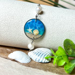 Elegantes Edelstahl-Armband mit rundem Resin-Anhänger in Meeresblau. Im Anhänger liegt echter Meersand als Ufersaum; darüber Sticker-Motive Seestern & Muschel. Zwei feine Süßwasserperlen verzieren das Armband. Verstellbarer Schiebeverschluss . Handgefertigtes Unikat – maritimer Schmuck, der an Wellenrauschen und Strandspaziergänge erinnert.