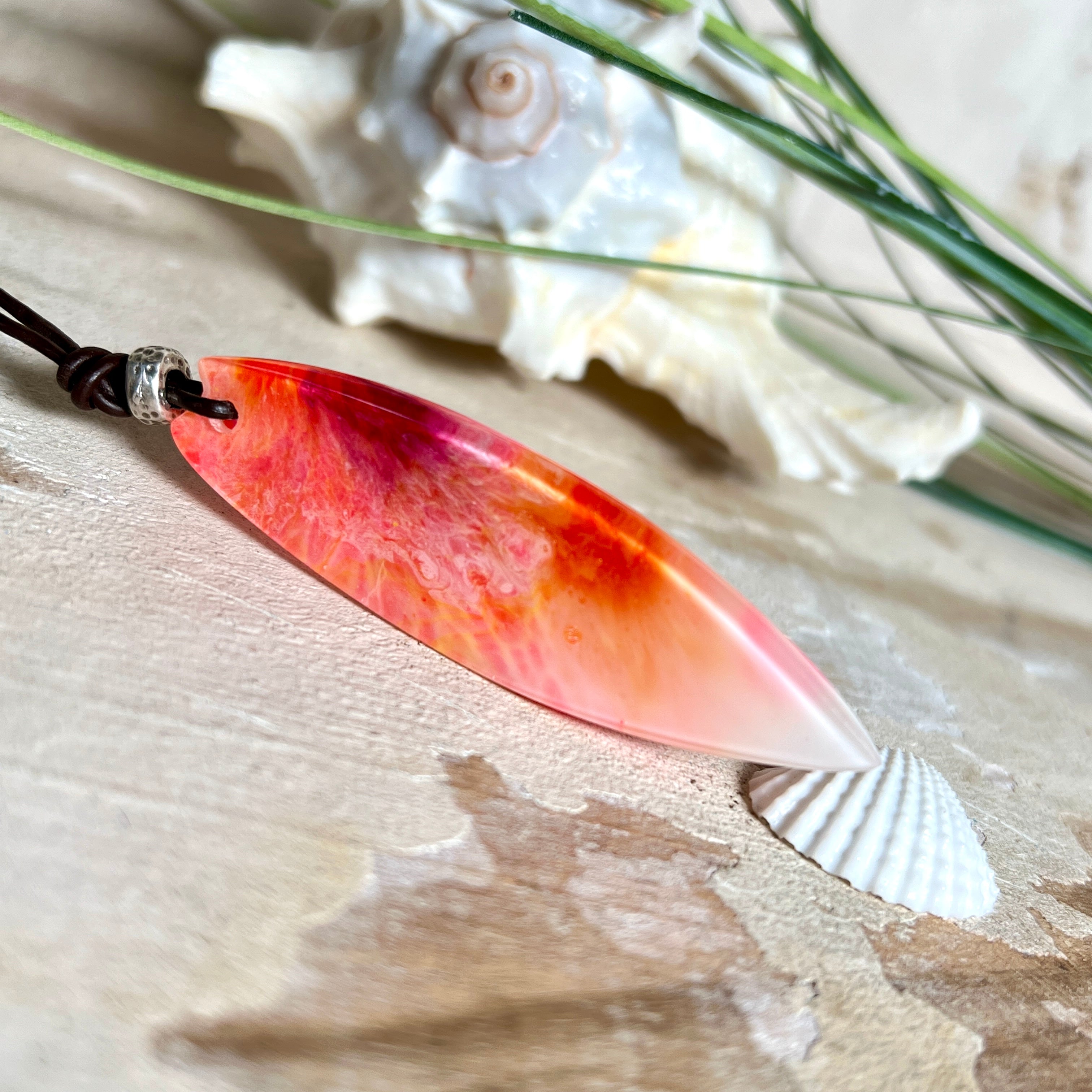 Handgefertigter Surfbrett-Anhänger aus fluoreszierendem Epoxidharz in leuchtenden Orange-, Rot- und Gelbtönen. Leuchtet bei Schwarzlicht. Kombiniert mit verstellbarem Lederband (26–45 cm), verziert mit Spacer- und Holzperlen. Das Design ist inspiriert von der Farbintensität eines Sonnenuntergangs über dem Meer.