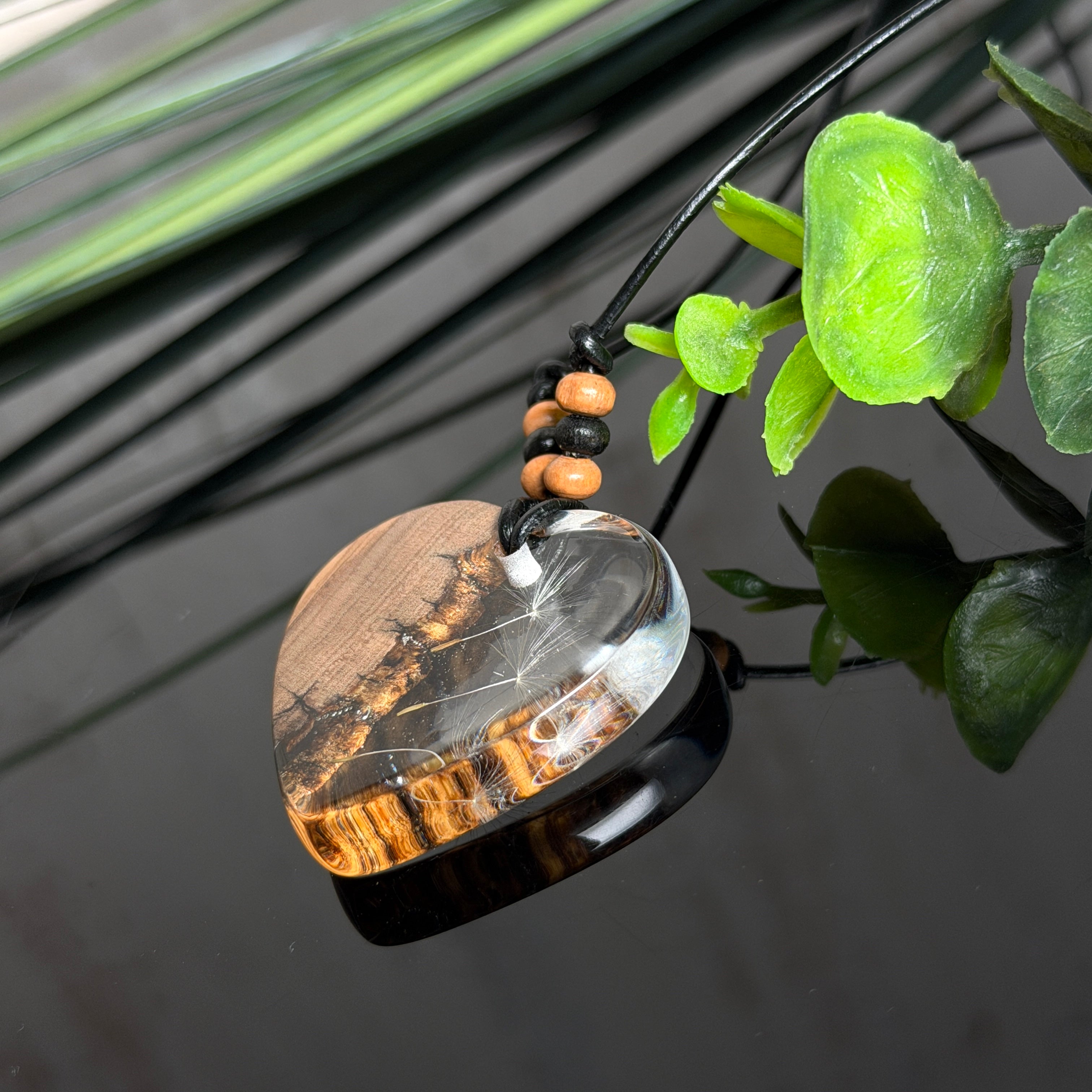 Handgefertigter Herzanhänger aus warm gemasertem Olivenholz und kristallklarem Epoxidharz mit schwebenden Pusteblumensamen. Schwarzes, verstellbares Lederband mit Schiebeknoten und naturfarbenen Holzperlen, Anhänger ca. 3,5 × 3,3 cm. Naturnaher Unikatschmuck von ZeitlosSchmuckDesign.