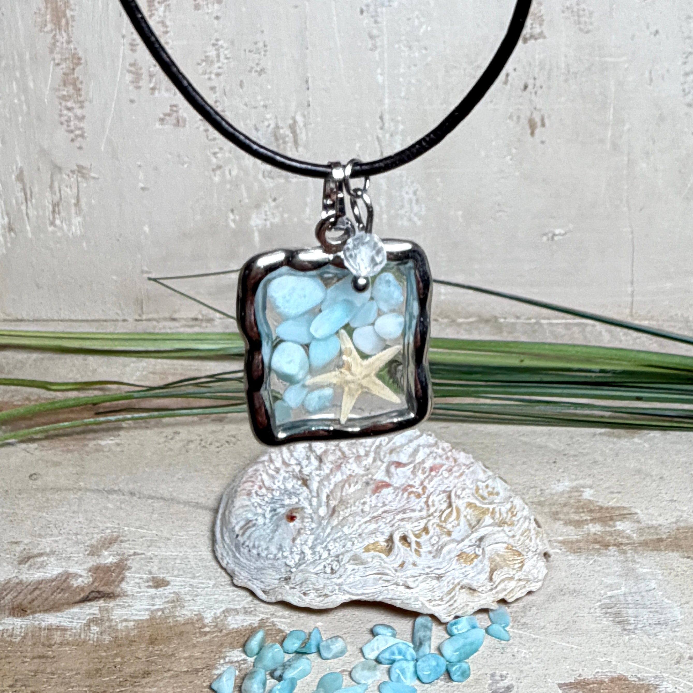 Quadratischer Larimar-Mosaik Anhänger im Edelstahlrahmen mit Seestern-Element und facettierter Bergkristall-Perle, eingebettet in Harz, an schwarzer Lederkette.