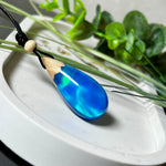 Handgefertigter Tropfenanhänger aus Maserholz und Harz mit synthetischem Opal. Die Farben schimmern je nach Lichteinfall in leuchtendem Blau und Grün – inspiriert von Polarlichtern. Der Anhänger ist ca. 3,9 × 1,8 cm groß und hängt an einem verstellbaren Lederband mit Holzperlen. Jedes Stück ist ein Unikat mit einzigartiger Holzmaserung. Ideal als Geschenk oder stilvoller Begleiter im Alltag.