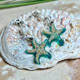 Handgefertigte Seestern-Ohrringe aus Epoxidharz und Sand mit Süßwasserperle, an 925 Sterling Silber Ohrsteckern; versilberte Anhängerschlaufe. Je Seestern ca. 2,6 × 2,5 cm. Leichte, maritime Dangle-Ohrringe im Strand-Look – ideal als Geschenk für Meeresliebhaberinnen.