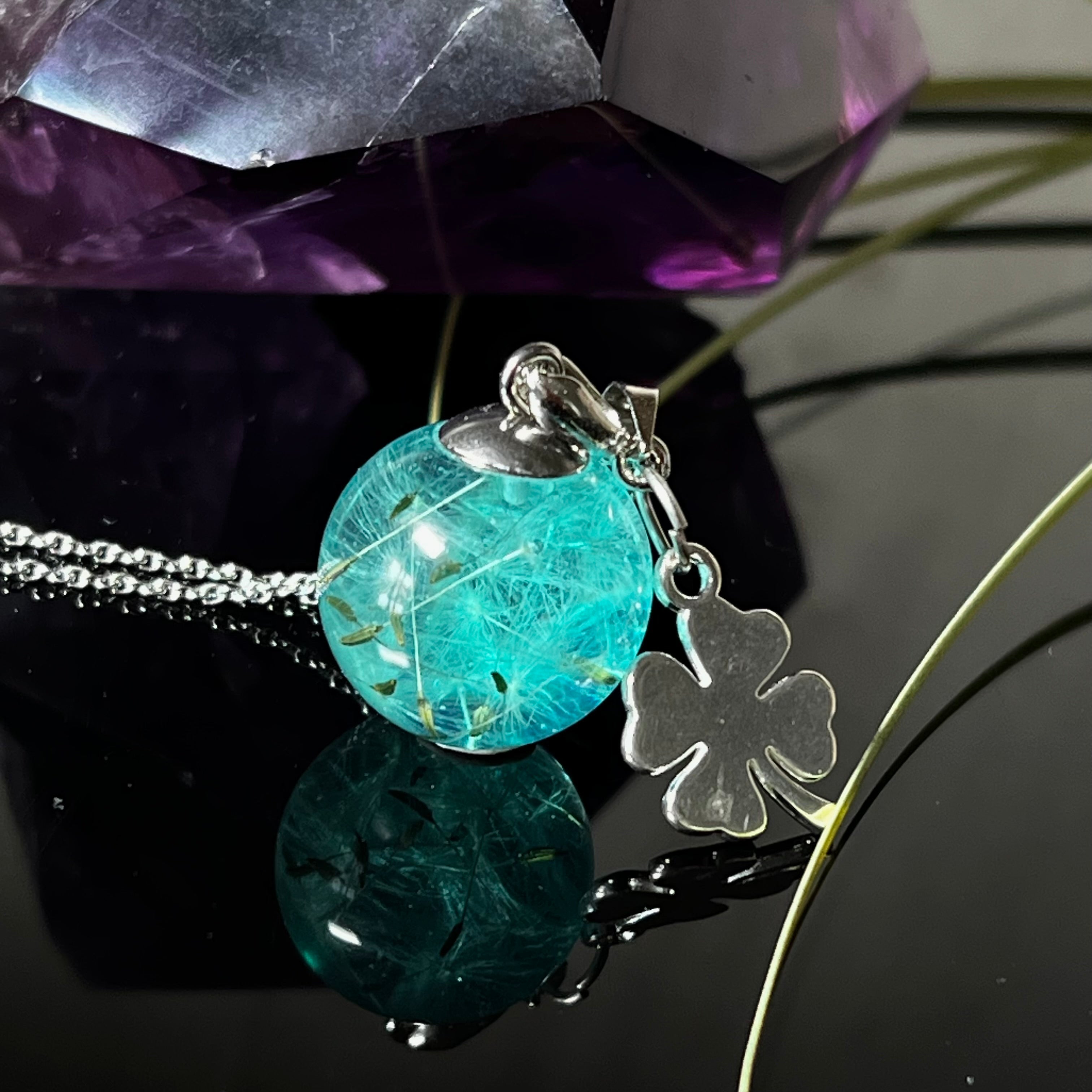 Eine handgefertigten Halskette mit türkiser Harzkugel, in der echte Pusteblumensamen eingeschlossen sind. Ergänzt durch einen glänzenden Edelstahl-Kleeblatt-Charm und einer feinen rhodinierten 925 Silberkette. Der Anhänger symbolisiert Glück, Leichtigkeit und neue Wege – ein emotionales Schmuckstück mit Bedeutung.