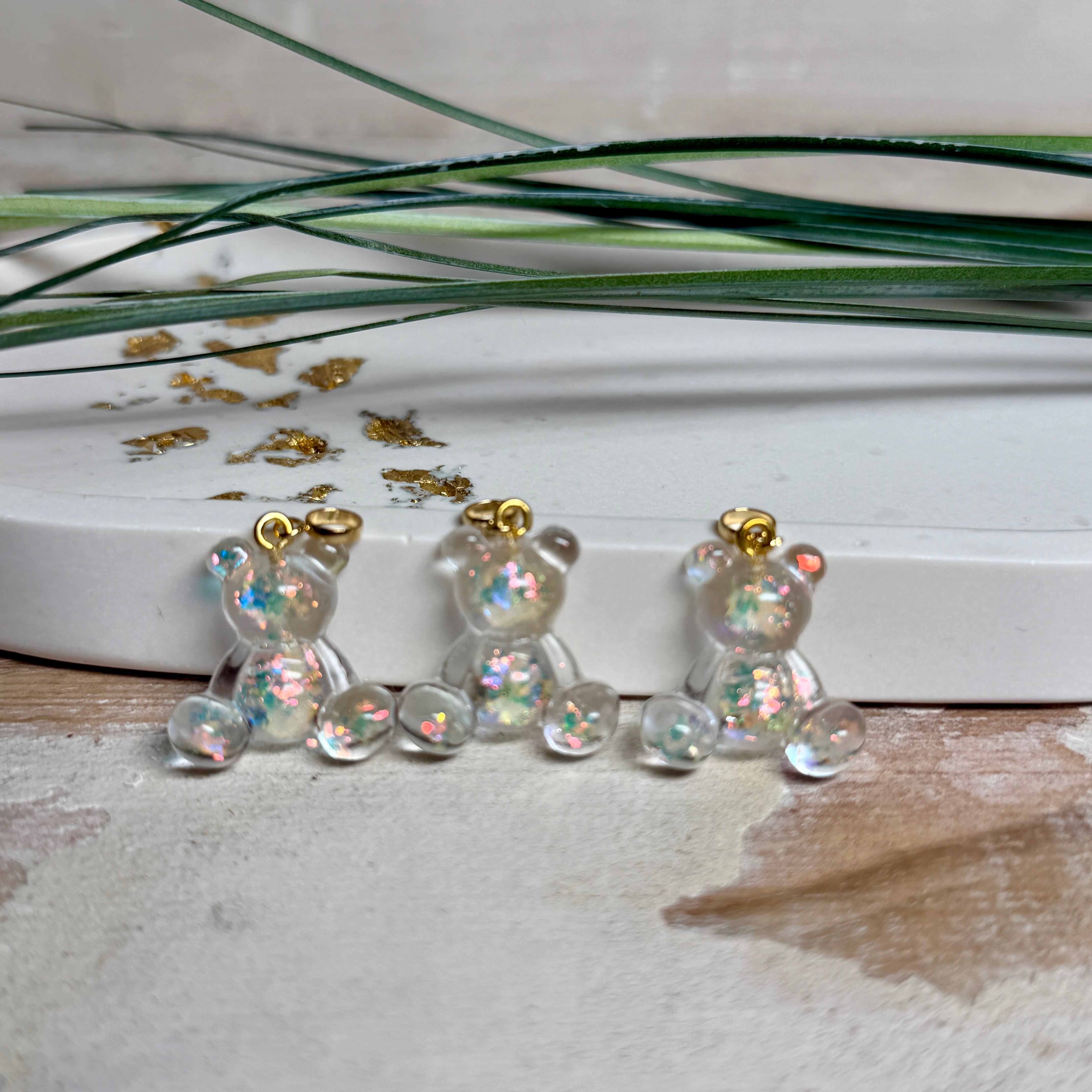 Kleiner Bärchen-Anhänger aus transparentem Harz mit irisierenden Flakes in Regenbogen-Schimmer, goldfarbene Anhängerschlaufe aus Edelstahl. Größe ca. 2,2 × 2,1 cm. Handgefertigtes Unikat; Flakes-Anordnung variiert, vereinzelt kleine Luftbläschen möglich.