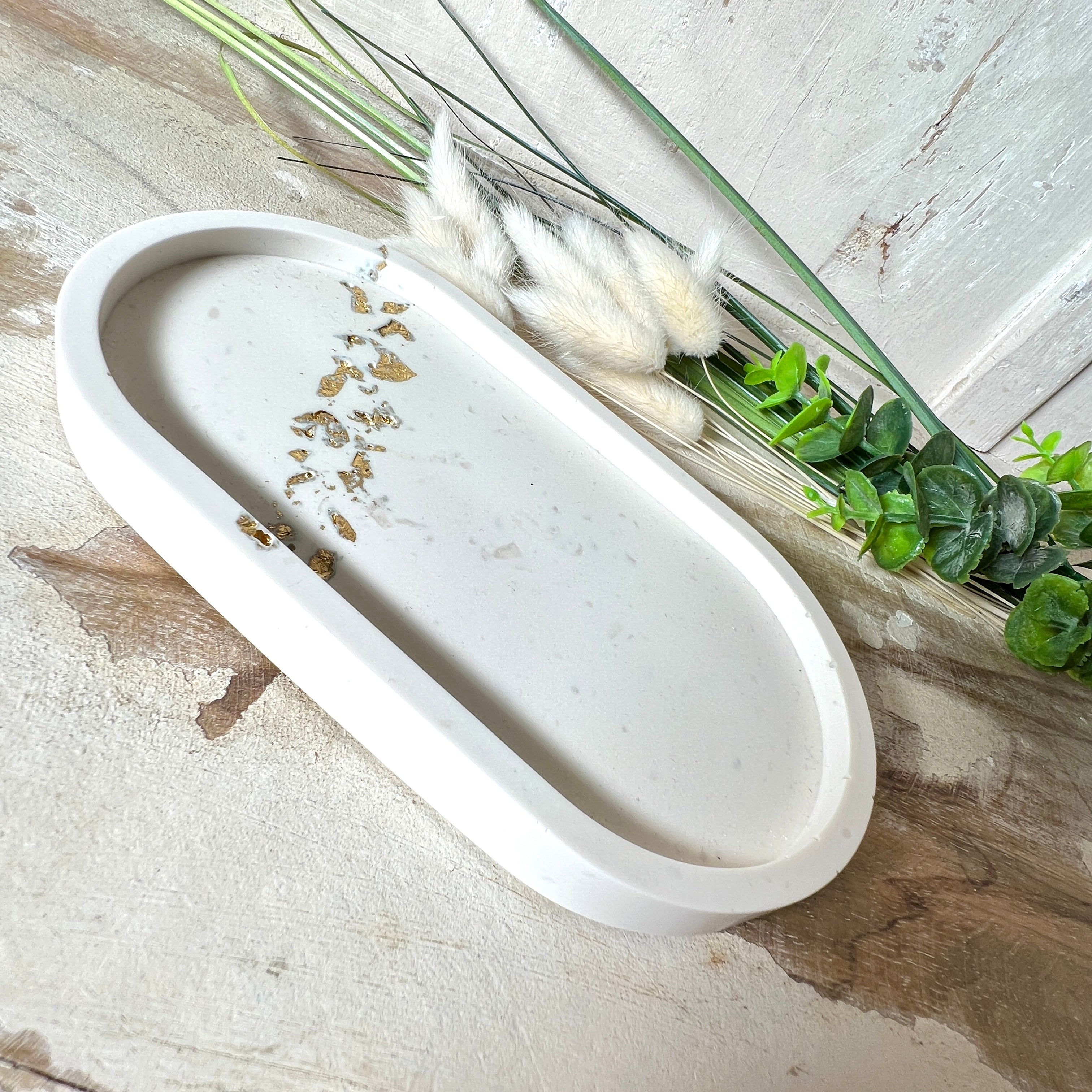 Handgefertigtes ovales Schmucktablett aus Jesmonite mit schimmernden Blattgold-Akzenten. Die Rückseite ist mit rutschfesten Korkpunkten ausgestattet. Ideal zur stilvollen Aufbewahrung von Schmuck, Schlüsseln oder kleinen Accessoires auf Kommoden und Nachttischen. Helles Design mit natürlicher Oberfläche und modernem Look. Unikat in feiner Handarbeit gefertigt – ohne Deko geliefert.