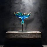 Handgefertigte Walflossen-Skulptur „Ocean Grace“ aus Maserholz und Epoxidharz in Blau, Weiß und Türkis, auf schwarzem Metallständer, ca. 29 x 27 cm, als exklusives maritimes Deko-Kunstwerk auf einem modernen Sideboard vor dunkler Wand platziert.