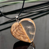 Pusteblumen Herzanhänger aus Olivenholz und glasklarem Epoxidharz, echte Löwenzahnsamen im Harz schwebend, schwarze Lederkette, handgefertigter Naturschmuck mit Spiegelung, dunkler Hintergrund