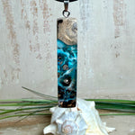 Rechteckiger Stabanhänger aus Maserholz und glasklarem Epoxidharz in tiefen Blau- und Türkistönen mit organischer Lagunen/Riff-Optik. Hochglanzpoliert, ca. 5,3 × 1,2 cm. Der Anhänger hängt an einer schwarzen Lederkette mit Edelstahl-Karabiner und 5-cm-Verlängerung (45 + 5 cm); die Anhängerschlaufe ist ebenfalls aus Edelstahl. Handgefertigtes Unikat, Lieferung in edler Schmuckbox – maritimer Look.