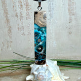 Rechteckiger Stabanhänger aus Maserholz und glasklarem Epoxidharz in tiefen Blau- und Türkistönen mit organischer Lagunen/Riff-Optik. Hochglanzpoliert, ca. 5,3 × 1,2 cm. Der Anhänger hängt an einer schwarzen Lederkette mit Edelstahl-Karabiner und 5-cm-Verlängerung (45 + 5 cm); die Anhängerschlaufe ist ebenfalls aus Edelstahl. Handgefertigtes Unikat, Lieferung in edler Schmuckbox – maritimer Look.