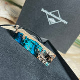 Rechteckiger Stabanhänger aus Maserholz und glasklarem Epoxidharz in tiefen Blau- und Türkistönen mit organischer Lagunen/Riff-Optik. Hochglanzpoliert, ca. 4,6 × 1,2 cm. Der Anhänger hängt an einer schwarzen Lederkette mit Edelstahl-Karabiner und 5-cm-Verlängerung (45 + 5 cm); die Anhängerschlaufe ist ebenfalls aus Edelstahl. Handgefertigtes Unikat, Lieferung in edler Schmuckbox – maritimer Look.