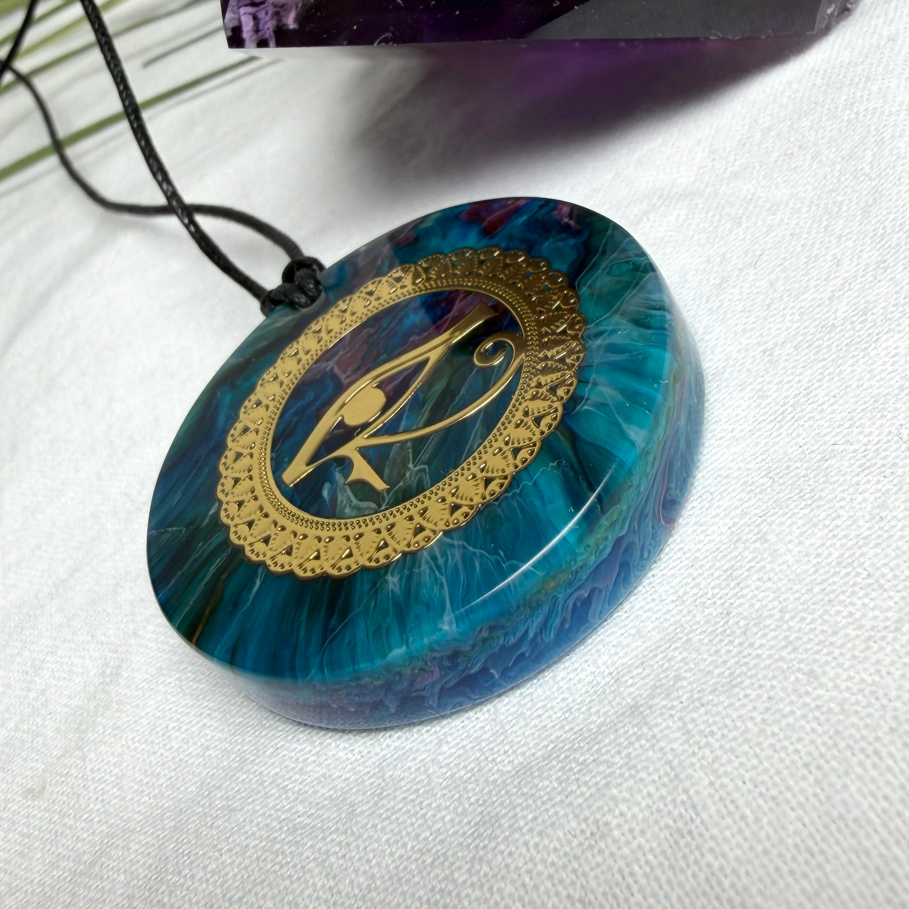 Runder Anhänger mit goldenem Horusauge aus Metall in blau türkis violettem Epoxidharz am schwarzen Baumwollband, spiritueller Eye of Ra Naturschmuck Amulett