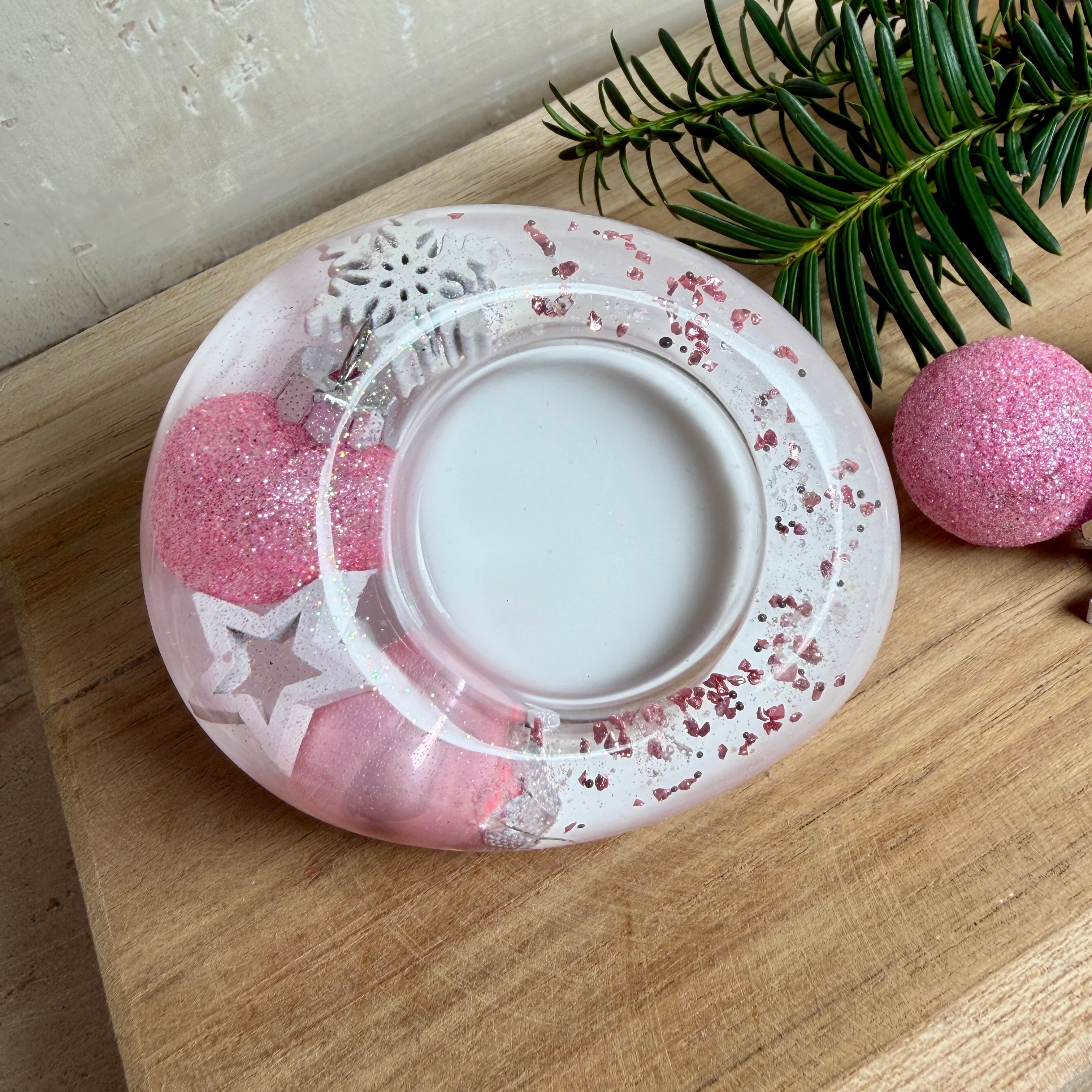 Rosa Teelichthalter aus klarem Epoxidharz, ca. 8 × 6,5 cm, Höhe ca. 4 cm. Im Harz sind kleine rosa Weihnachtskugeln, ein Stern, eine Schneeflocke und rosa Granulat eingegossen. Ovaler Halter für ein Teelicht, Unterseite mit vier runden Korkpads als Möbelschoner. Fotografiert auf Holz mit Tannenzweig und rosa Kugeln als weihnachtlicher Dekohintergrund.