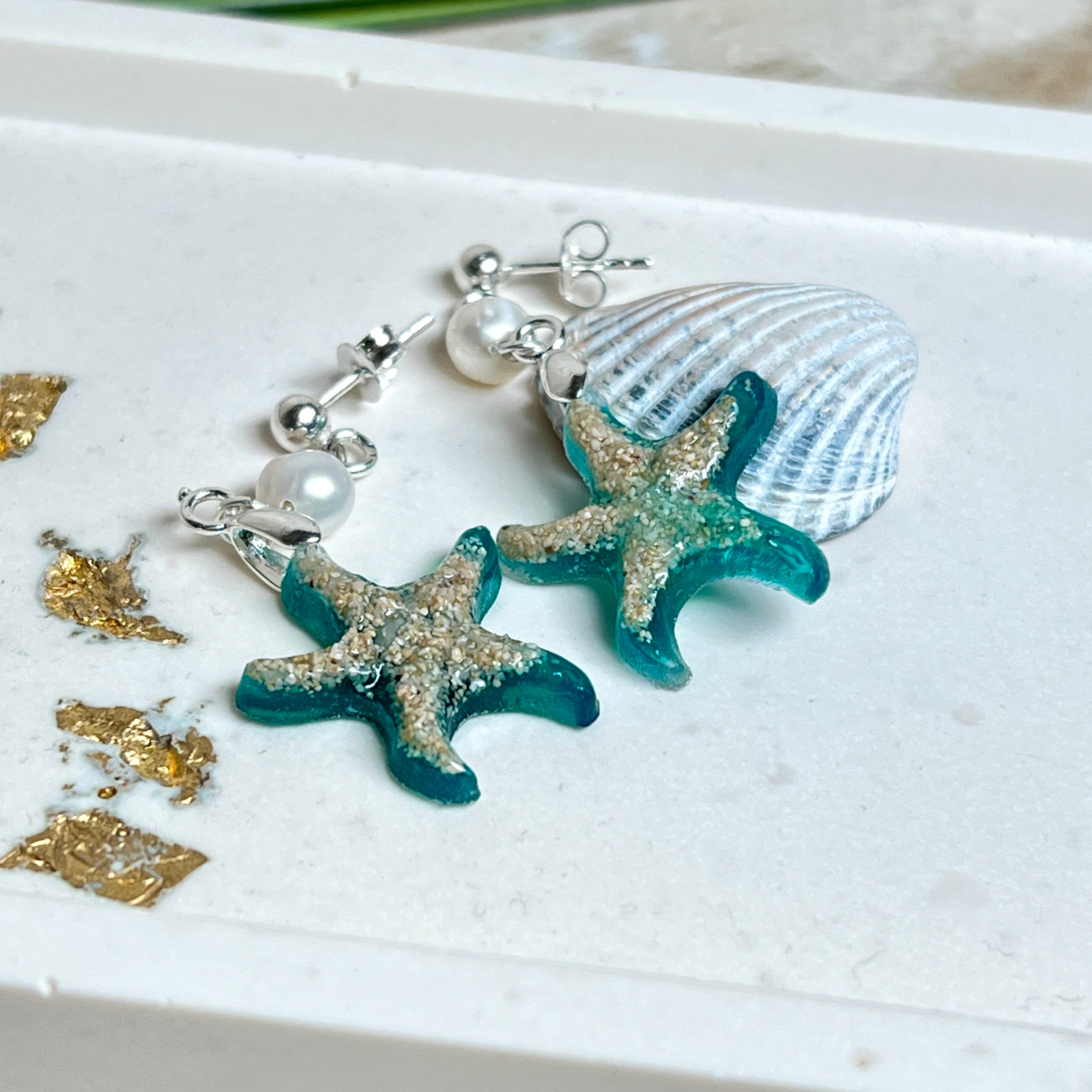 Handgefertigte Seestern-Ohrringe aus Epoxidharz und Sand mit Süßwasserperle, an 925 Sterling Silber Ohrsteckern; versilberte Anhängerschlaufe. Je Seestern ca. 2,6 × 2,5 cm. Leichte, maritime Dangle-Ohrringe im Strand-Look – ideal als Geschenk für Meeresliebhaberinnen.