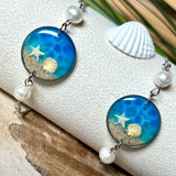 Elegantes Edelstahl-Armband mit rundem Resin-Anhänger in Meeresblau. Im Anhänger liegt echter Meersand als Ufersaum; darüber Sticker-Motive Seestern & Muschel. Zwei feine Süßwasserperlen verzieren das Armband. Verstellbarer Schiebeverschluss . Handgefertigtes Unikat – maritimer Schmuck, der an Wellenrauschen und Strandspaziergänge erinnert.