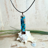 Rechteckiger Stabanhänger aus Maserholz und glasklarem Epoxidharz in tiefen Blau- und Türkistönen mit organischer Lagunen/Riff-Optik. Hochglanzpoliert, ca. 5,3 × 1,2 cm. Der Anhänger hängt an einer schwarzen Lederkette mit Edelstahl-Karabiner und 5-cm-Verlängerung (45 + 5 cm); die Anhängerschlaufe ist ebenfalls aus Edelstahl. Handgefertigtes Unikat, Lieferung in edler Schmuckbox – maritimer Look.