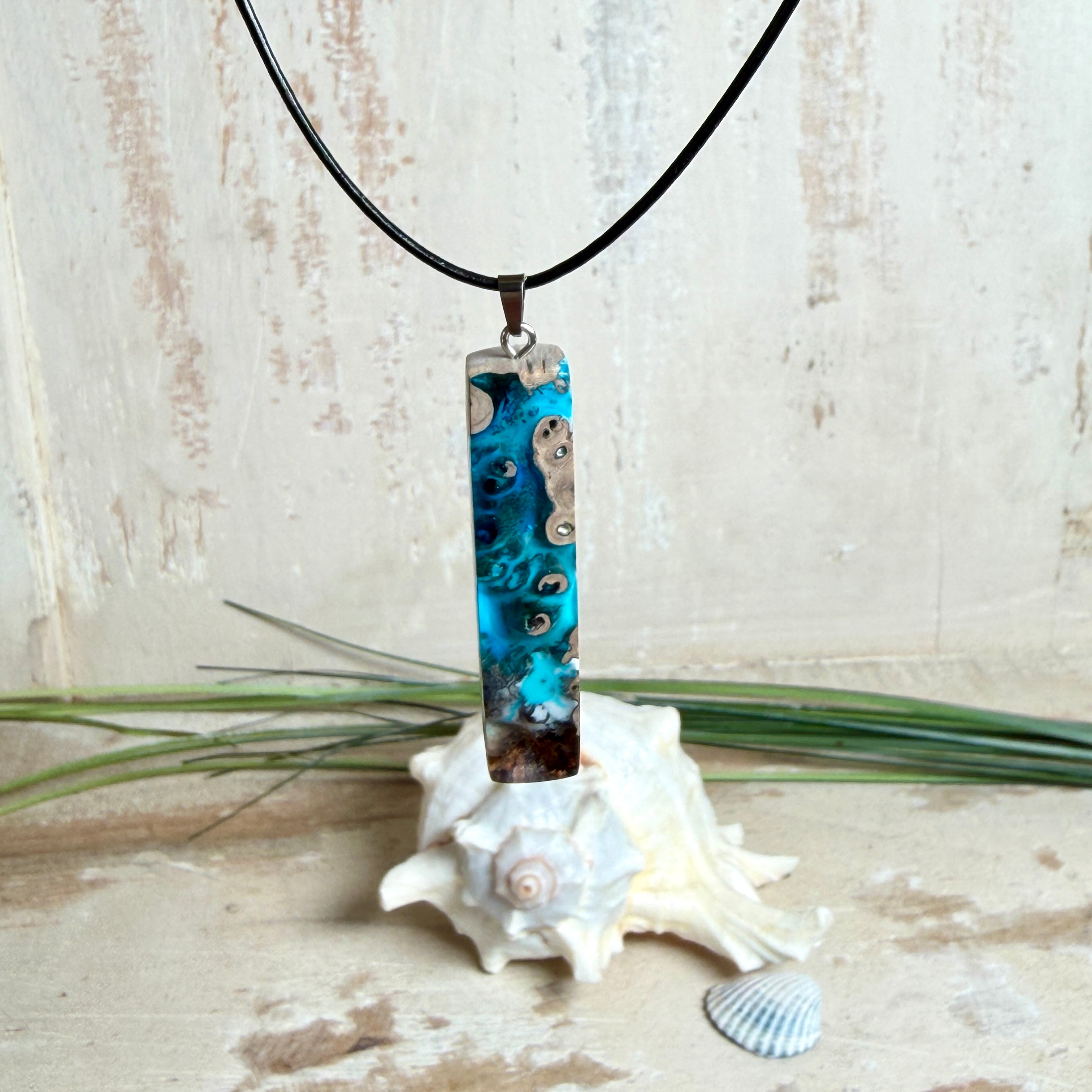 Rechteckiger Stabanhänger aus Maserholz und glasklarem Epoxidharz in tiefen Blau- und Türkistönen mit organischer Lagunen/Riff-Optik. Hochglanzpoliert, ca. 5,3 × 1,2 cm. Der Anhänger hängt an einer schwarzen Lederkette mit Edelstahl-Karabiner und 5-cm-Verlängerung (45 + 5 cm); die Anhängerschlaufe ist ebenfalls aus Edelstahl. Handgefertigtes Unikat, Lieferung in edler Schmuckbox – maritimer Look.