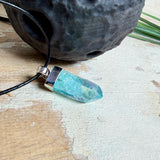 Nahaufnahme einer handgefertigten Halskette mit Aqua Aura Rohkristall: naturbelassener, sechskantiger Bergkristall mit goldbedampfter Aqua-Beschichtung in Türkisblau, gefasst in 925er Silber und getragen an einer schwarzen Lederkette mit Edelstahl-Karabiner und Verlängerungskettchen.