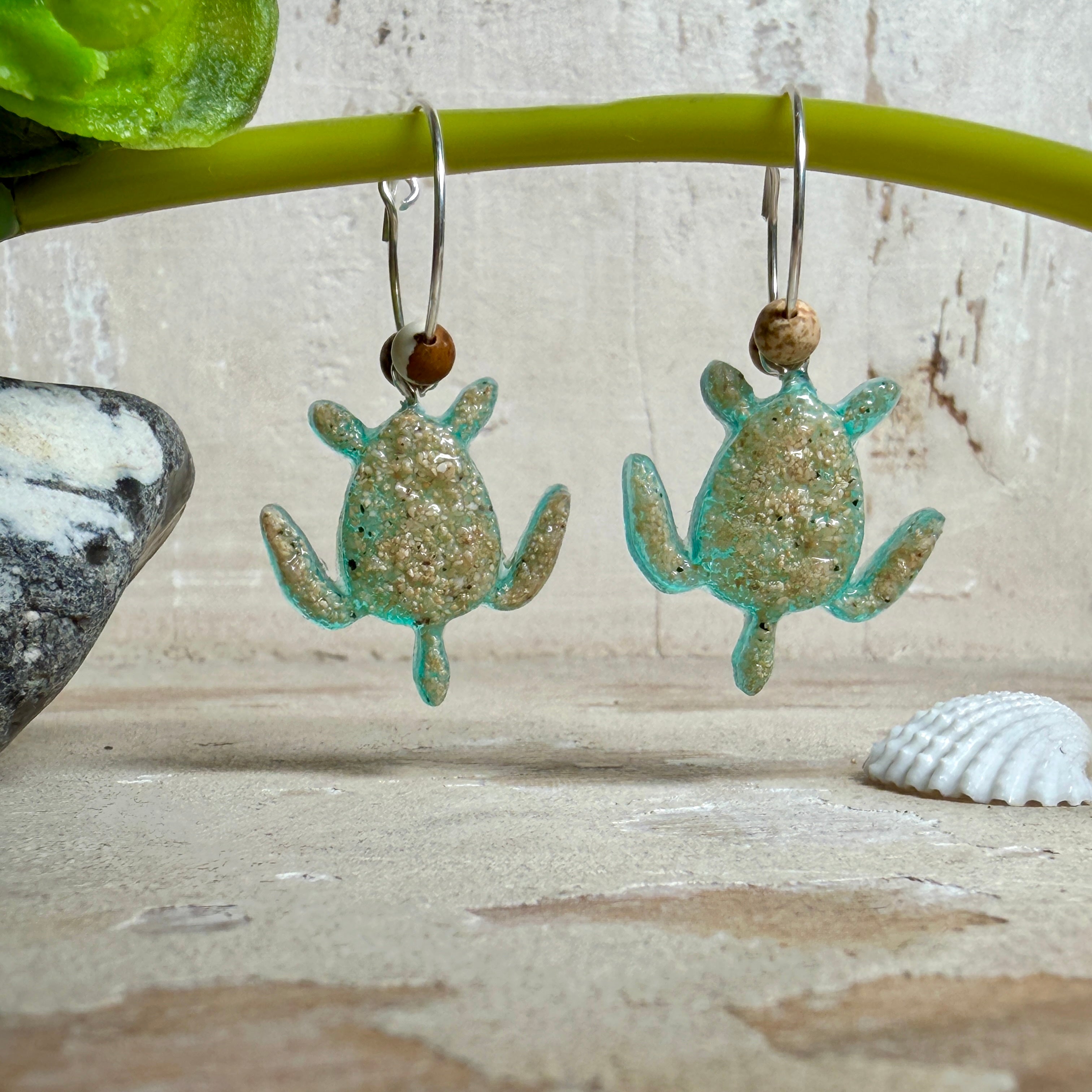 Türkisfarbene Schildkröten-Creolen an feinen 16-mm-Ringen aus 925 Silber, jeweils mit echtem Strandsand im Harz eingebettet und mit kleinen Jaspis-Edelsteinperlen verziert; handgefertigtes Unikat mit sommerlich-maritimem Look, fotografiert als Freisteller, Nahaufnahme oder auf dekorativem Untergrund.