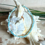 Zartes Larimar-Armband „Lagunenlicht“ mit facettierten 3 mm Perlen aus seltenem Atlantisstein auf Elastikfaden. Die kleinen Larimarsteine zeigen sanfte Blau- und Weißtöne wie Lagunenwasser. Innenumfang ca. 19 cm. Fotografiert auf Muscheln, einem dekorativen Stein und in einer schwarzen Schmuckbox von ZeitlosSchmuckDesign im maritimen Stil.
