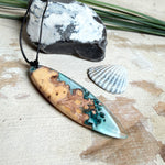 Handgefertigter, länglicher Surfbrett-Anhänger aus naturbelassenem Maserholz und transparentem Harz in Lagunen-Türkis, an schwarzem Lederband mit Knoten – maritimer Unikatschmuck für Damen und Herren, dekorativ mit Stein und Muschel inszeniert.