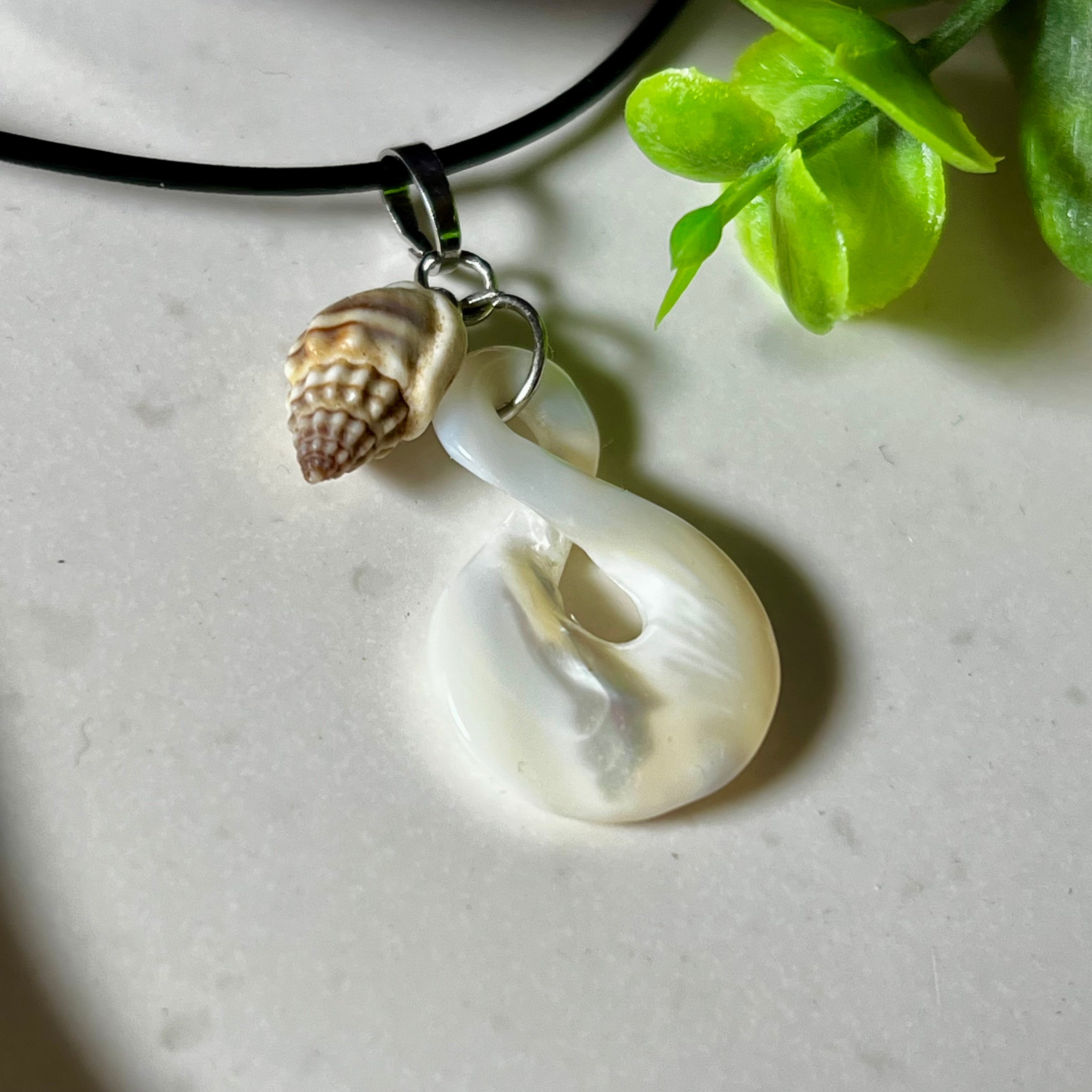 Detailaufnahme eines handgefertigten Anhängers aus echtem Perlmutt in Schlaufenform, kombiniert mit einer kleinen echten Muschel als Charm-Anhänger. Der Schmuck hängt an einer feinen schwarzen Lederschnur mit Edelstahlverschluss und symbolisiert Harmonie und Unendlichkeit. Ideal für Meeresliebhaberinnen und Fans von Naturmaterialien.