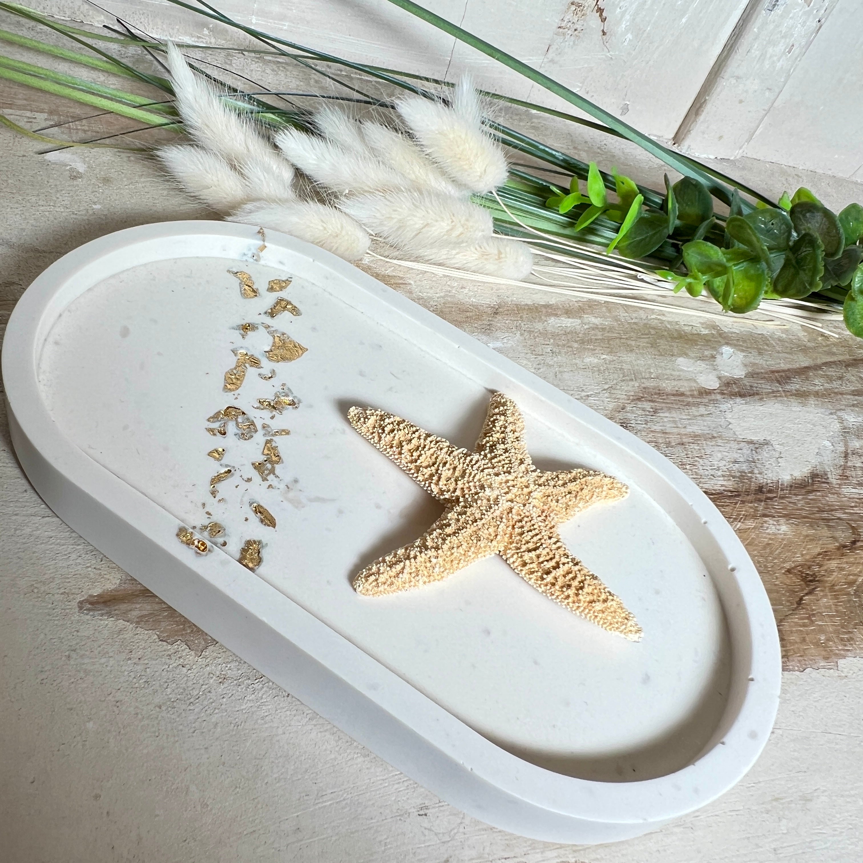 Handgefertigtes ovales Schmucktablett aus Jesmonite mit schimmernden Blattgold-Akzenten. Die Rückseite ist mit rutschfesten Korkpunkten ausgestattet. Ideal zur stilvollen Aufbewahrung von Schmuck, Schlüsseln oder kleinen Accessoires auf Kommoden und Nachttischen. Helles Design mit natürlicher Oberfläche und modernem Look. Unikat in feiner Handarbeit gefertigt – ohne Deko geliefert.