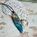 Unikat Anhänger aus stabilisiertem Holz mit türkisblauem Harzverlauf, schlanke Form, poliert. Verstellbares braunes Lederband 22–46 cm mit Schiebeknoten. Anhänger ca. 6,6 × 2 cm. Tragefoto am Hals und Produktfotos mit maritimer Deko