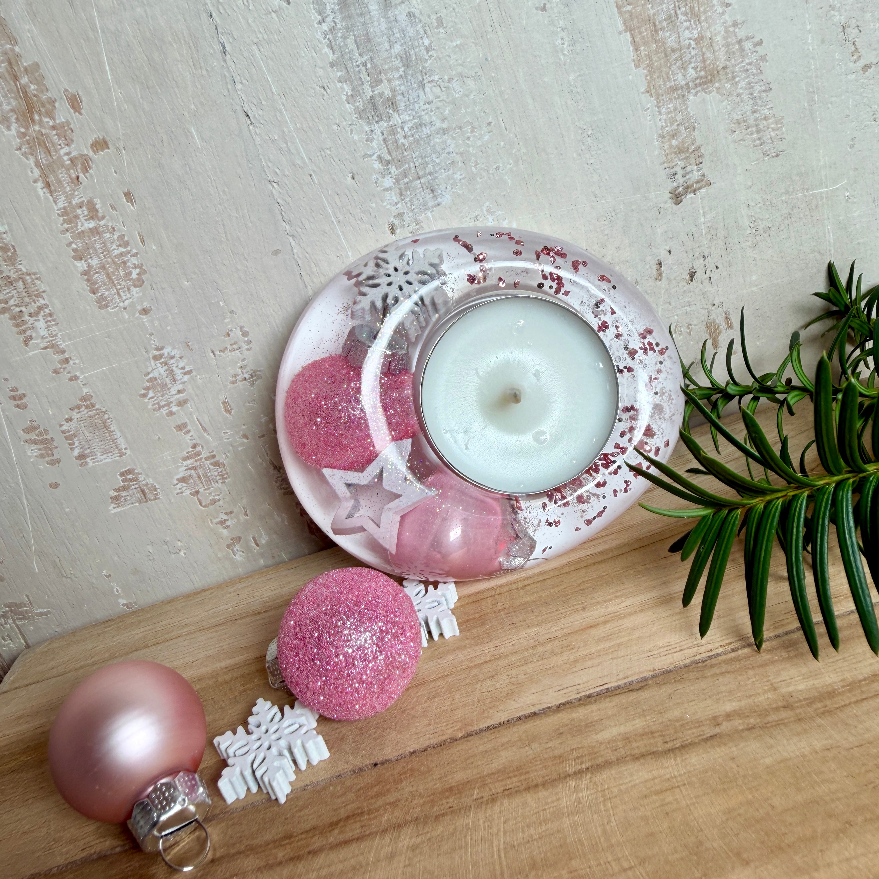 Rosa Teelichthalter aus klarem Epoxidharz, ca. 8 × 6,5 cm, Höhe ca. 4 cm. Im Harz sind kleine rosa Weihnachtskugeln, ein Stern, eine Schneeflocke und rosa Granulat eingegossen. Ovaler Halter für ein Teelicht, Unterseite mit vier runden Korkpads als Möbelschoner. Fotografiert auf Holz mit Tannenzweig und rosa Kugeln als weihnachtlicher Dekohintergrund.