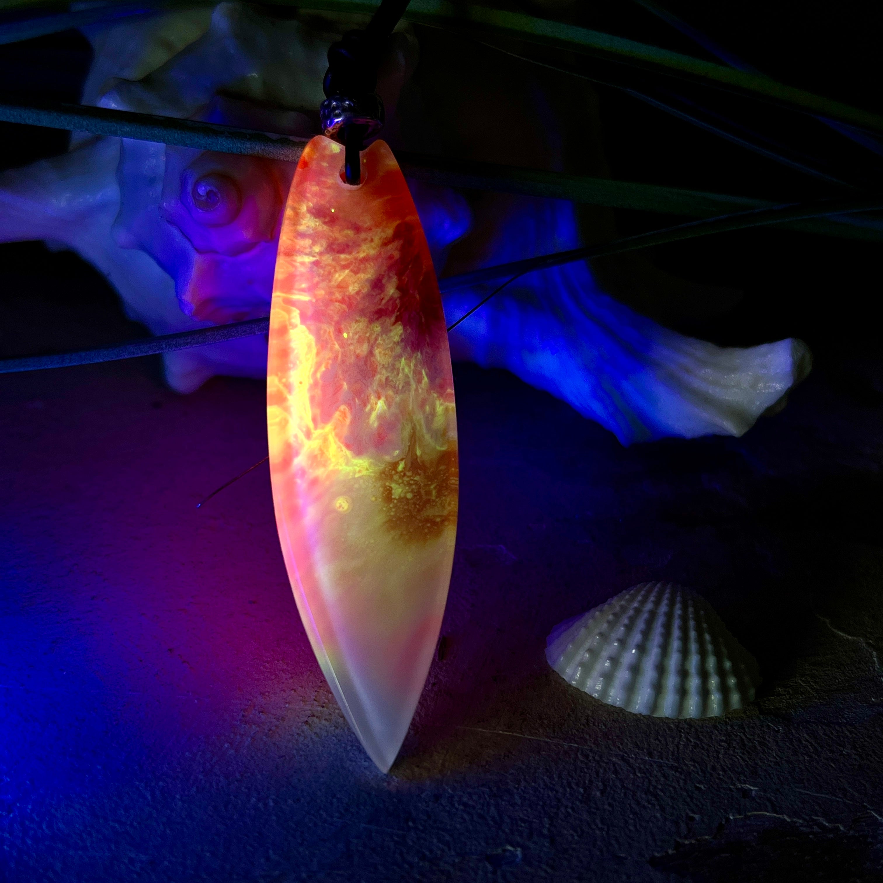 Handgefertigter Surfbrett-Anhänger aus fluoreszierendem Epoxidharz in leuchtenden Orange-, Rot- und Gelbtönen. Leuchtet bei Schwarzlicht. Kombiniert mit verstellbarem Lederband (26–45 cm), verziert mit Spacer- und Holzperlen. Das Design ist inspiriert von der Farbintensität eines Sonnenuntergangs über dem Meer.