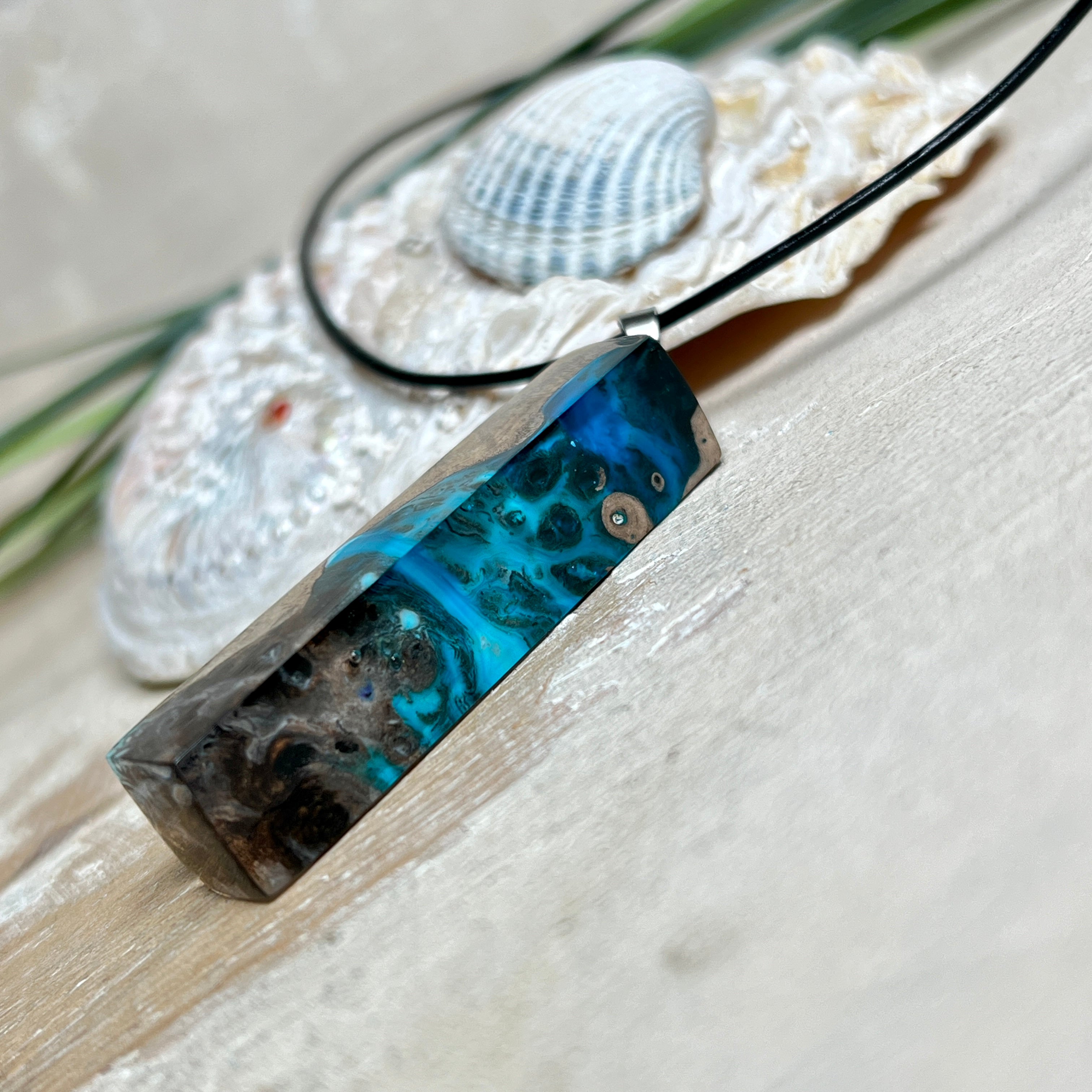 Rechteckiger Stabanhänger aus Maserholz und glasklarem Epoxidharz in tiefen Blau- und Türkistönen mit organischer Lagunen/Riff-Optik. Hochglanzpoliert, ca. 4,6 × 1,2 cm. Der Anhänger hängt an einer schwarzen Lederkette mit Edelstahl-Karabiner und 5-cm-Verlängerung (45 + 5 cm); die Anhängerschlaufe ist ebenfalls aus Edelstahl. Handgefertigtes Unikat, Lieferung in edler Schmuckbox – maritimer Look.