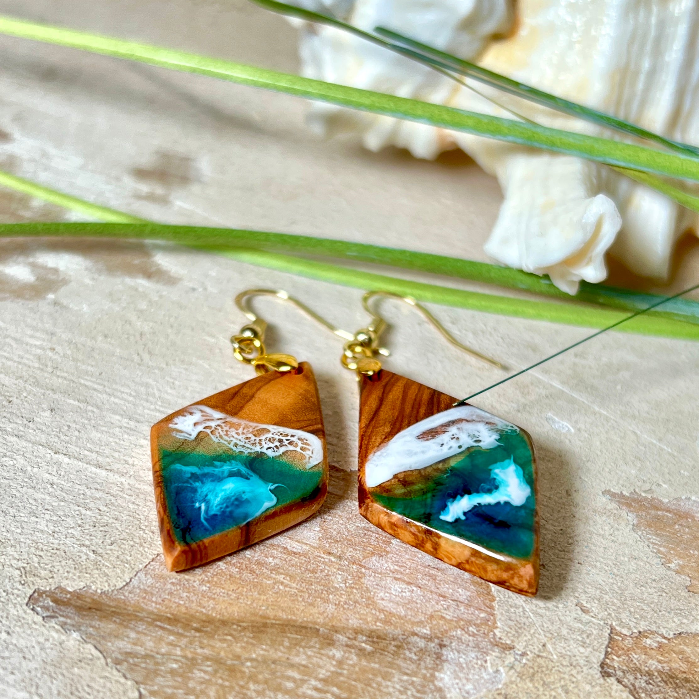 Handgefertigte Ohrringe „Ocean Diamond“ aus Olivenholz und Epoxidharz in Diamantform. Türkis-, Blau- und Weißtöne bilden ein wellenartiges Design, inspiriert vom Meer. Jeder Ohrring ist ein Unikat mit individueller Maserung. Vergoldete, nickelfreie und bleifreie Anhängerschlaufe, 925 Sterling Silber Ohrhaken. Maritimer Schmuck für Meerliebhaber und besondere Anlässe.