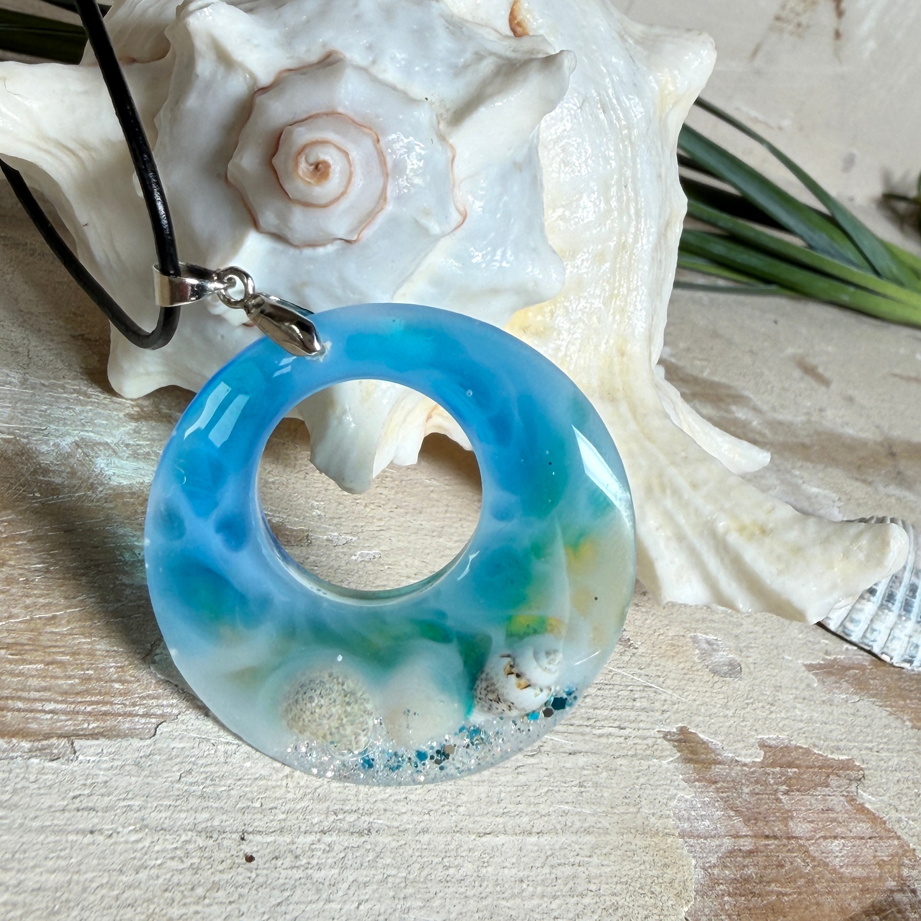 Runder Harzanhänger „Ocean Circle“ mit 4 cm Durchmesser in Blau-Weiß-Verlauf, in dem kleine Muscheln und feiner Glitzer wie ein Mini-Strand eingefangen sind. Der Anhänger hat eine Edelstahl-Schlaufe und hängt an einer schwarzen Lederkette mit Edelstahl-Karabiner und Verlängerung. Maritimer, handgefertigter Unikat-Schmuck im Lagunen-Look für Meeresliebhaberinnen.
