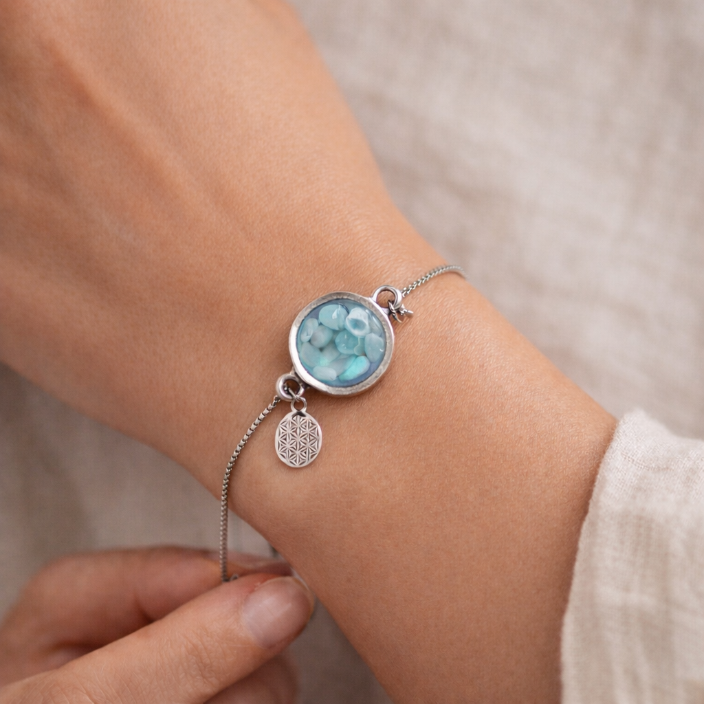Verstellbares Edelstahlarmband mit Zirkoniasteinchen und Schiebeverschluss und rundem Anhänger (ca. 2,5 × 1,7 cm) mit echten Larimar-Edelstein-Splittern in Harz. Zusätzlich ein Charm „Blume des Lebens“ aus 925 Silber. Handgefertigtes Unikat in Türkis- und Meeresblautönen – Schmuckstück in Nahaufnahme bzw. als Tragebild am Handgelenk.