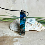 Rechteckiger Stabanhänger aus Maserholz und glasklarem Epoxidharz in tiefen Blau- und Türkistönen mit organischer Lagunen/Riff-Optik. Hochglanzpoliert, ca. 4,6 × 1,2 cm. Der Anhänger hängt an einer schwarzen Lederkette mit Edelstahl-Karabiner und 5-cm-Verlängerung (45 + 5 cm); die Anhängerschlaufe ist ebenfalls aus Edelstahl. Handgefertigtes Unikat, Lieferung in edler Schmuckbox – maritimer Look.