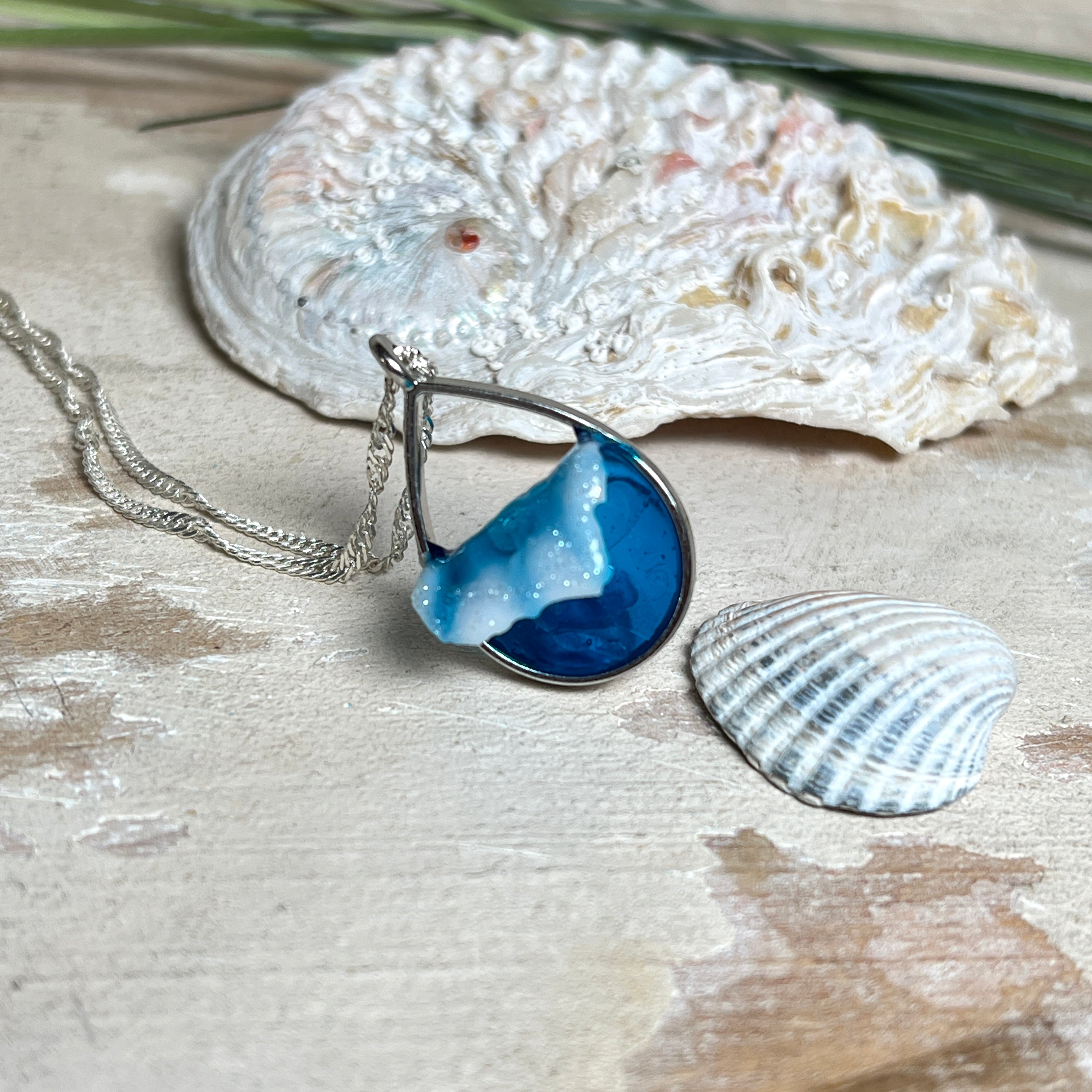 „Lagunenlicht“ – Tropfenanhänger mit handmodellierter 3D-Welle und feinem Glitzereffekt. Klarer Harzlook in Blau/Türkis, gefasst in Silber-Legierung. Größe ca. 3,5 × 2 cm. Geliefert an einer versilberten Kette (ca. 56 cm). Hochglänzend , maritimer Unikatcharakter.