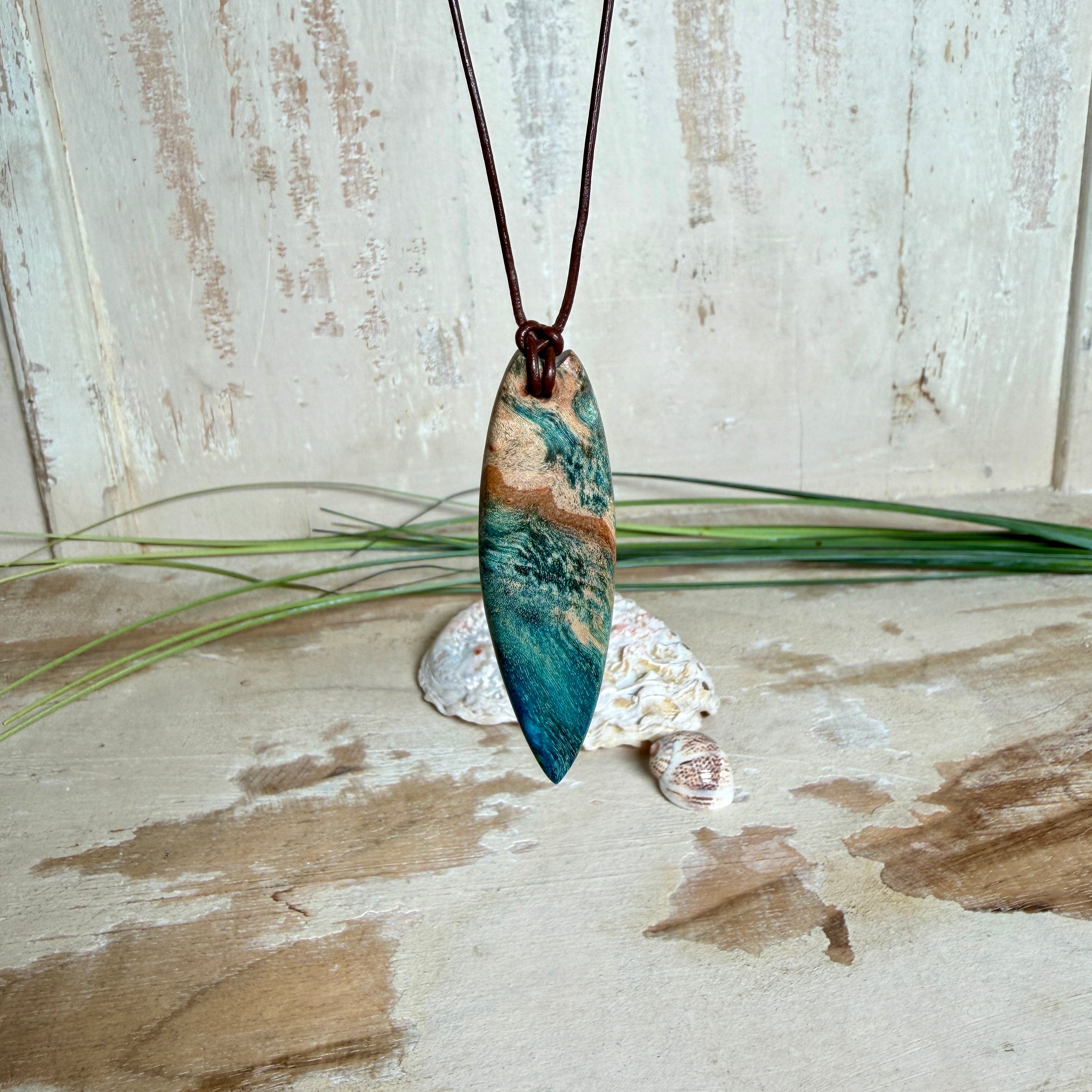 Unikat Anhänger aus stabilisiertem Holz mit türkisblauem Harzverlauf, schlanke Form, poliert. Verstellbares braunes Lederband 22–46 cm mit Schiebeknoten. Anhänger ca. 6,6 × 2 cm. Tragefoto am Hals und Produktfotos mit maritimer Deko