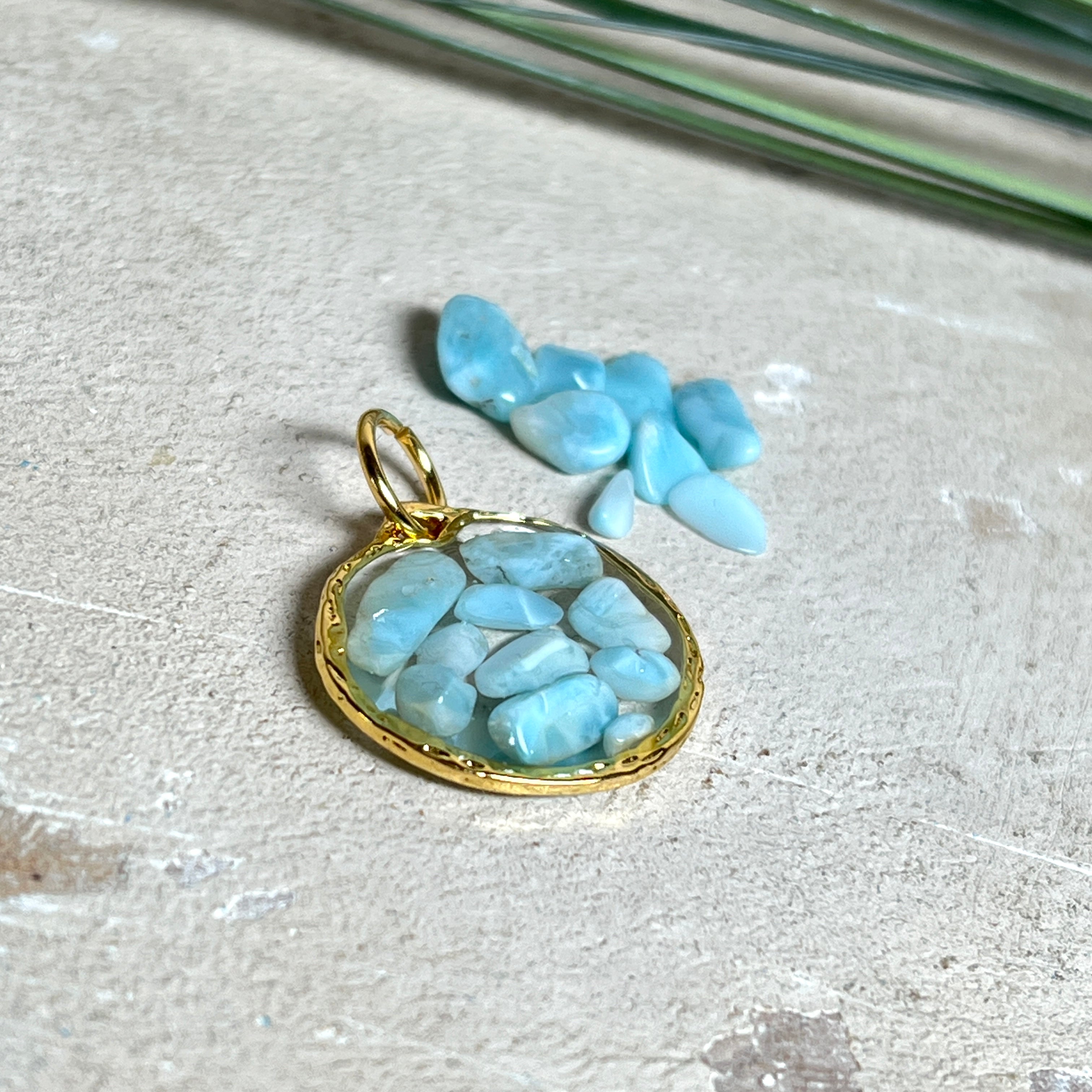Runder Anhänger „Golden Atlantis Dream“ mit kleinen Larimar-Edelsteinchen in klarem Resin, gefasst in einer 24K vergoldeten Zinklegierung in Shiny Gold. Die Larimarsteine zeigen sanfte Blau- und Weißtöne wie karibische Wellen. Durchmesser ca. 22 mm, handgefertigtes Unikat, fotografiert mit Larimarsteinchen, Muscheln und einer 2-Euro-Münze zur Größenorientierung auf maritimem Untergrund.