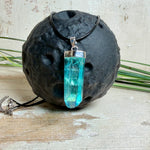 Nahaufnahme einer handgefertigten Halskette mit Aqua Aura Rohkristall: naturbelassener, sechskantiger Bergkristall mit goldbedampfter Aqua-Beschichtung in Türkisblau, gefasst in 925er Silber und getragen an einer schwarzen Lederkette mit Edelstahl-Karabiner und Verlängerungskettchen.