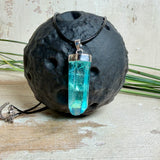 Nahaufnahme einer handgefertigten Halskette mit Aqua Aura Rohkristall: naturbelassener, sechskantiger Bergkristall mit goldbedampfter Aqua-Beschichtung in Türkisblau, gefasst in 925er Silber und getragen an einer schwarzen Lederkette mit Edelstahl-Karabiner und Verlängerungskettchen.