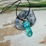 Nahaufnahme einer handgefertigten Halskette mit Aqua Aura Rohkristall: naturbelassener, sechskantiger Bergkristall mit goldbedampfter Aqua-Beschichtung in Türkisblau, gefasst in 925er Silber und getragen an einer schwarzen Lederkette mit Edelstahl-Karabiner und Verlängerungskettchen.